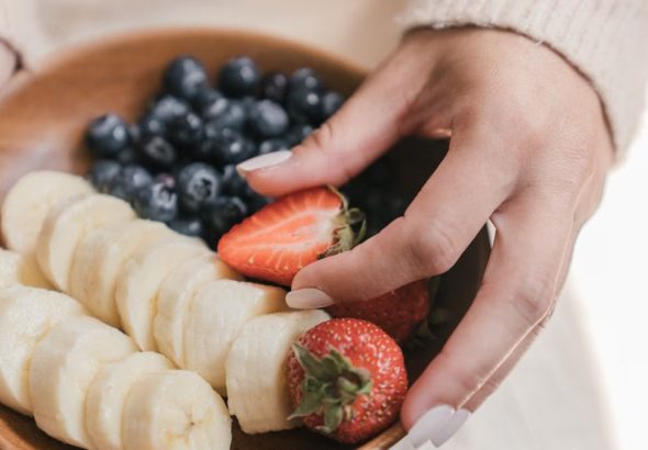 A wooden bowl filled with fresh bananas and berries held by an unrecognizable person's hands.
